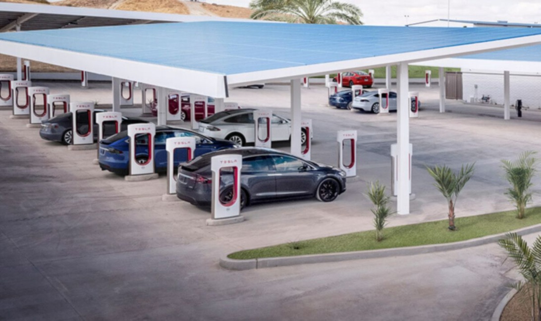 En Tesla Supercharger-station med flera laddplatser under ett blått solcellstak. Flera Tesla-elbilar är parkerade och anslutna till de distinkta röda och vita laddenheterna i en ökenliknande miljö med palmer.